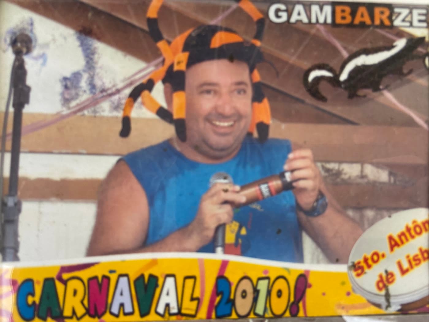 Feijão sorrindo ao microfone no Carnaval 2010, com um chapéu de aranha listrado de laranja e preto, embaixo do letreiro da Gambarzeira e do gambá pintado