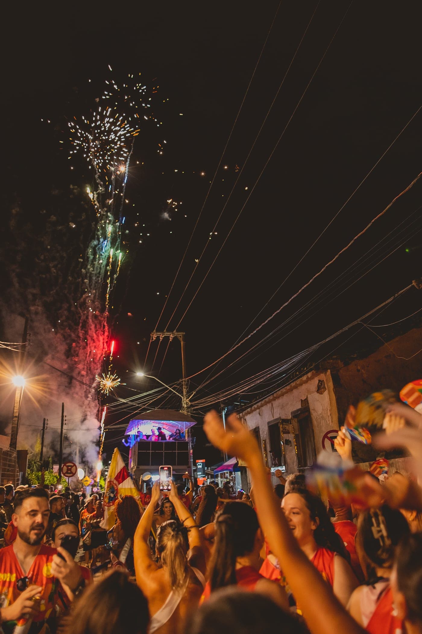 Fogos de artifício sobre o trio do bloco, com a multidão de braços erguidos na rua do casario