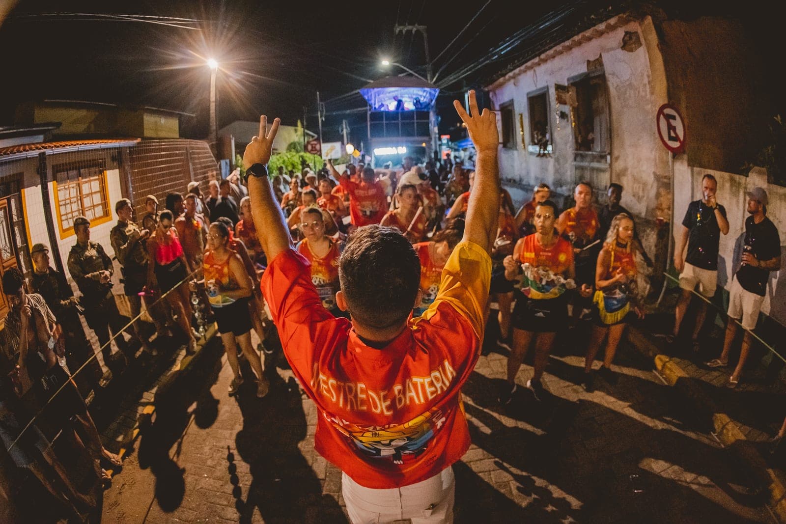 O mestre de bateria de costas, braços erguidos, regendo a bateria na rua estreita da Freguesia