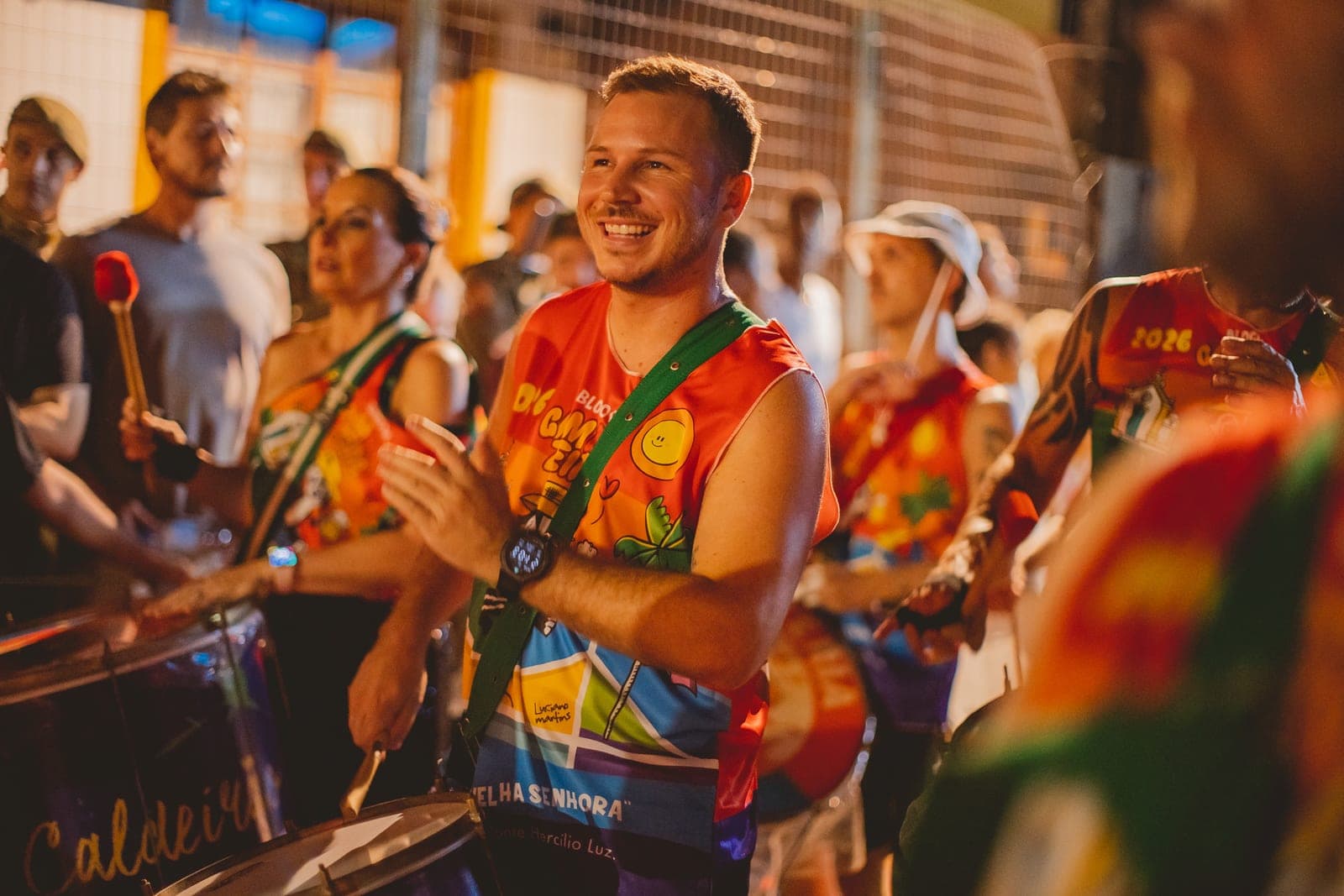 Ritmista sorrindo no meio da bateria, com a camiseta do bloco ilustrada por Luciano Martins